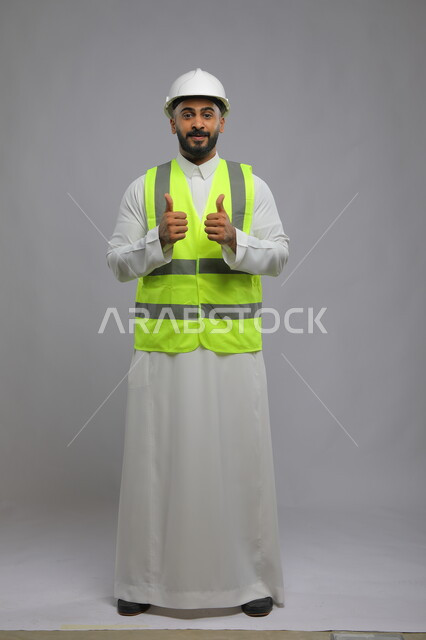 Following up on the progress of projects, architectural construction in Saudi Arabia, art in design and construction technology, a full-length portrait of a smiling Saudi Arabian Gulf engineer wearing a helmet and protection vest, raising his thumbs upwards with movements of admiration and acceptance, precision and skill, gray background.