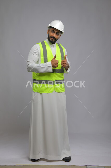 الإنشاءات الإعمارية في الأراضي السعودية ،مفهوم الهندسة والانشاءات المعمارية، جودة الفن في تقنية التصميم والبناء، بورتريه لمهندس عربي خليجي سعودي مبتسم يرتدي الخوذه و سترة الحماية يرفع الابهام للأعلى بحركات الاعجاب والقبول، صورة الجسم بالكامل، خلفية رمادية