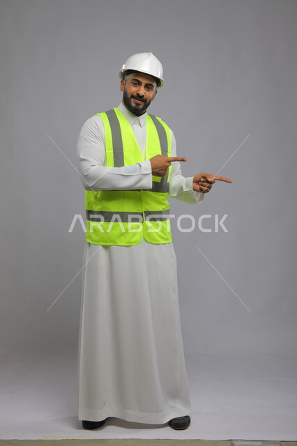 مهن ووظائف سعودية، تطور وتقدم المباني على يد مهندسين سعوديين ،مفهوم الانشاءات المعمارية والهندسة، بورتريه لمهندس عربي خليجي سعودي مبتسم يرتدي الخوذه و سترة الحماية يشير بأصابعه إلى موقع العمل، صورة الجسم  بالكامل ، خلفية رمادية