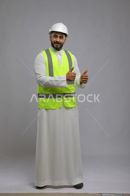 The development and progress of buildings at the hands of Saudi engineers, the concept of architectural construction and engineering, a portrait of a smiling Saudi Arabian Gulf engineer wearing a helmet and protection vest, raising a thumbs up with gestures of admiration and acceptance and the quality mark, Saudi professions and jobs, full-body portrait, gray background