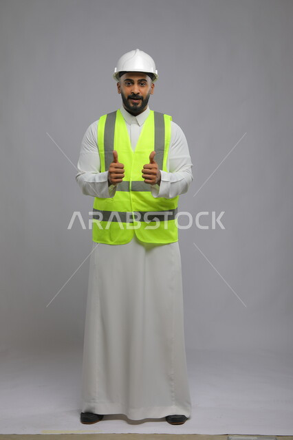The concept of architectural and engineering constructions, the quality of art in design and construction technology, work in the engineering sector, a portrait of a smiling Saudi Arabian Gulf engineer wearing a helmet and protection vest, raising a thumbs up with gestures of admiration and acceptance, Saudi professions and jobs, full-body portrait, gray background
