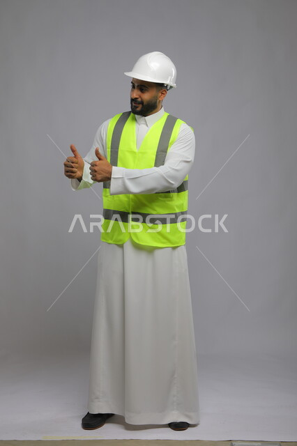 جودة الفن في تقنية التصميم والبناء، العمل في القطاع الهندسي، مفهوم الهندسة والانشاءات المعمارية، بورتريه لمهندس عربي خليجي سعودي مبتسم يرتدي الخوذه و سترة الحماية يرفع الابهام للأعلى بحركات الاعجاب والقبول، مهن ووظائف سعودية، صورة لكامل الجسم، خلفية رمادية