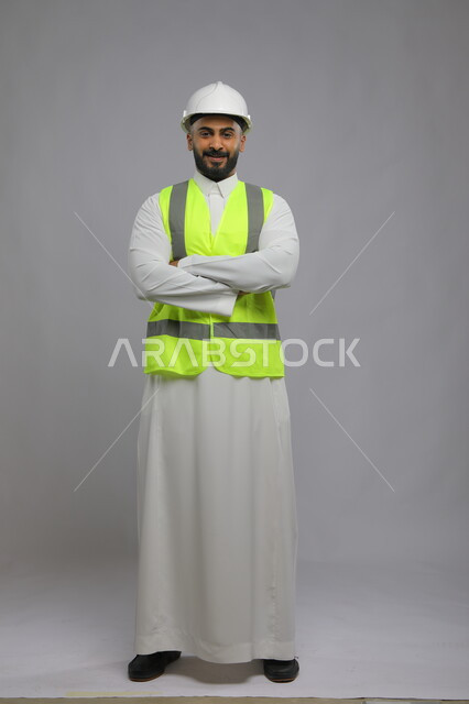  العمل في القطاع الهندسي، مفهوم الهندسة والانشاءات المعمارية، بورتريه لمهندس عربي خليجي سعودي مبتسم يرتدي الخوذه و سترة الحماية ينظر إلى الكاميرا، الوقوف بإيماءات أيدي متقاطعه، مهن ووظائف سعودية، الإعمار بسواعد أبناء الوطن، صورة لكامل الجسم، خلفية رمادية