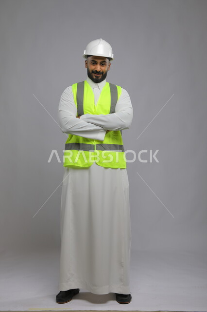 مفهوم الهندسة والانشاءات المعمارية، بورتريه لمهندس عربي خليجي سعودي مبتسم يرتدي الخوذه و سترة الحماية   ينظر إلى الكاميرا، الوقوف بإيماءات أيدي متقاطعه، العمل في القطاع الهندسي، مهن ووظائف سعودية، الإعمار بسواعد أبناء الوطن، صورة لكامل الجسم، خلفية رمادية