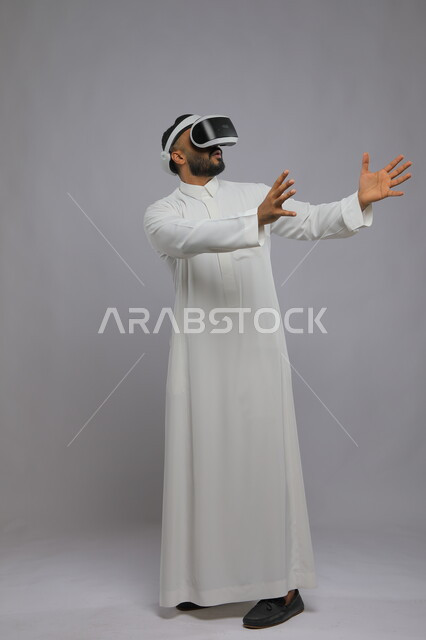 الاندماج والتفاعل مع ألعاب الواقع الافتراضي, التقنيات والأجهزة المتطورة, محاكاة العالم الخيالي ومحاولة لمس عنصر ما, بورتريه لرجل عربي سعودي خليجي يرتدي  الزي التقليدي ونظارة في آر ثلاثية الأبعاد الذكية, صورة الجسم بالطول الكامل, خلفية رمادية.