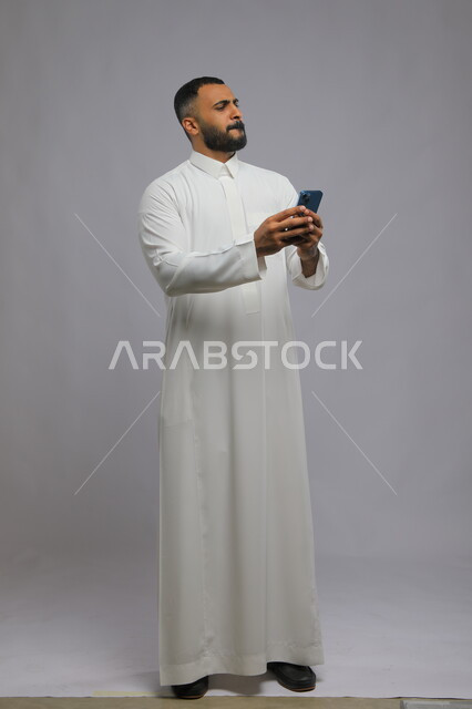 ارسال رسالة نصية عبر الجوال, ايماءات الحزن والانزعاج لخسارة صفقة عمل, بورتريه لرجل عربي خليجي سعودي يرتدي الثوب التقليدي يقوم باستخدام الهاتف المحمول, صورة طول الجسم الكامل, خلفية رمادية