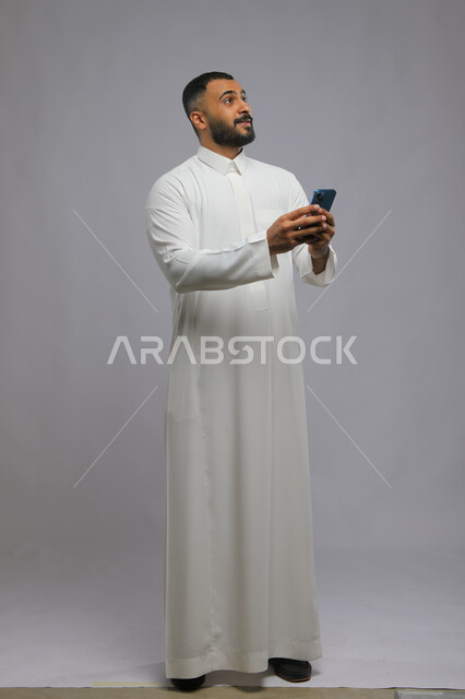 النظر إلى شيء ما بإيماءات التمني, مفهوم شرود الذهن والتأمل والتفكير, بورتريه لشاب عربي خليجي سعودي يرتدي الثوب التقليدي يقوم باستخدام الهاتف المحمول, التواصل والدردشات مع الأهل والأصدقاء عبر الانترنت, صورة الجسم بالطول الكامل, خلفية رمادية