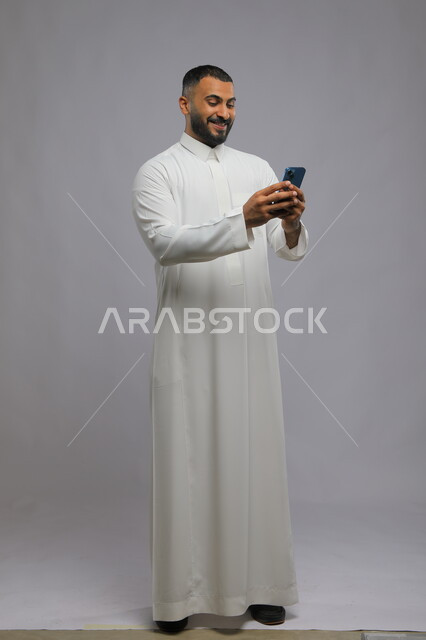 التسويق الالكتروني عبر الأجهزة الذكية, تصفح مواقع التواصل الاجتماعي بإيماءات السرور, متابعة آخر أخبار  السوشال ميديا, بورتريه لرجل عربي خليجي سعودي يرتدي الثوب التقليدي يستخدم الهاتف المحمول, صورة الجسم بالطول الكامل, خلفية رمادية