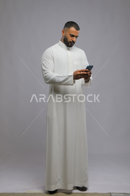 ارسال رسالة نصية عبر الجوال, ايماءات الحزن والانزعاج لخسارة صفقة عمل, بورتريه لرجل عربي خليجي سعودي يرتدي الثوب التقليدي يقوم باستخدام الهاتف المحمول, صورة طول الجسم الكامل, خلفية رمادية