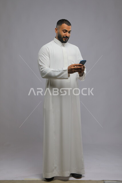 التسويق الالكتروني عبر الأجهزة الذكية, تصفح مواقع التواصل الاجتماعي بإيماءات السرور, متابعة آخر أخبار  السوشال ميديا, بورتريه لرجل عربي خليجي سعودي يرتدي الثوب التقليدي يستخدم الهاتف المحمول, صورة الجسم بالطول الكامل, خلفية رمادية