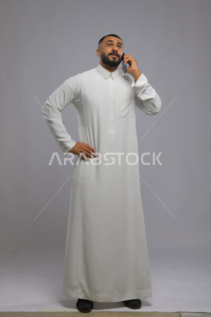 دردشات وتعارف باستخدام الهاتف المحمول, التواصل مع الأصدقاء بحب وود, بورتريه لرجل عربي خليجي سعودي يرتدي الثوب التقليدي يقوم بإجراء مكالمة هاتفية عبر الجوال, صورة الجسم بالكامل, خلفية رمادية