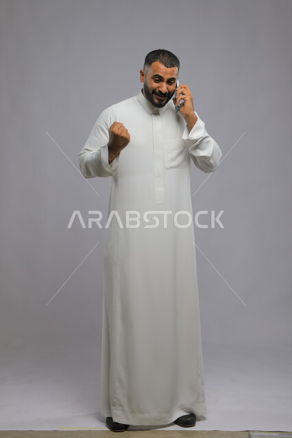 إجراء مكالمة هاتفية, إيماءات السعادة والسرور لسماع الأخبار الجيدة, الدردشة والتحدث مع الأصدقاء عبر الجوال, بورتريه لشاب عربي خليجي سعودي يرتدي الثوب التقليدي يقوم بالرد على اتصال الهاتف المحمول, صورة الجسم بالكامل, خلفية رمادية