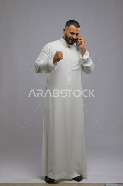 إجراء مكالمة هاتفية, إيماءات السعادة والسرور لسماع الأخبار الجيدة, الدردشة والتحدث مع الأصدقاء عبر الجوال, بورتريه لشاب عربي خليجي سعودي يرتدي الثوب التقليدي يقوم بالرد على اتصال الهاتف المحمول, صورة الجسم بالكامل, خلفية رمادية