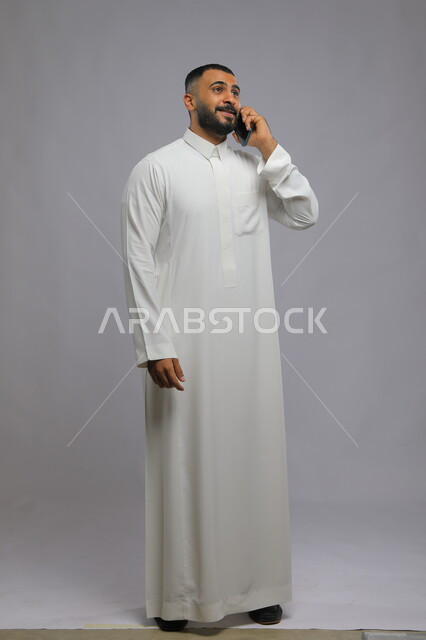 التواصل مع زملاء العمل باستخدام الهاتف المحمول, إيمات الفرح بتحقيق الأهداف وزيادة الأرباح, بورتريه لرجل عربي خليجي سعودي يرتدي الثوب التقليدي يقوم بإجراء مكالمة هاتفية عبر الجوال, صورة الجسم بالكامل, خلفية رمادية