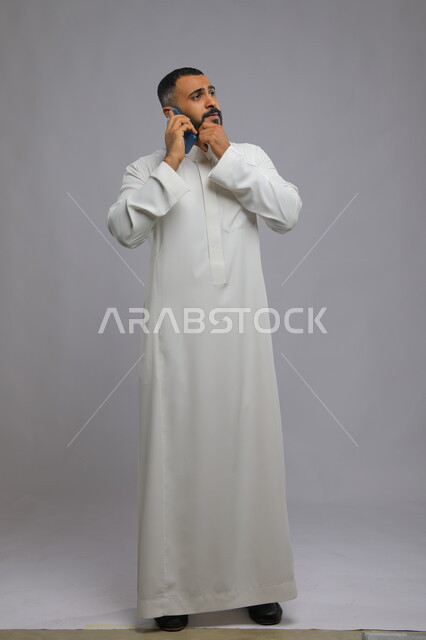 التفكير لإيجاد حل لمشكلة ما, الشعور بالانزعاج لسماع الاخبار السيئة, ايماءات الحيرة والاستفهام, بورتريه لرجل عربي خليجي سعودي يرتدي الثوب التقليدي يقوم بإجراء مكالمة هاتفيةعبر الجوال, دردشات ومحادثات مع الأصدقاء, صورة الجسم بالكامل, خلفية رمادية
