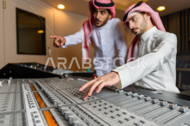 شابان سعوديان في غرفة خارج الاستديو ،يتم معالجةالاصوات المسجلة داخل الاستديو عن طريق برمجيات صوتية من خلال جهاز الحاسوب المخصص ، استديو تسجيل صوتي ، بث مباشر ، فريق الاعداد ، الاذاعة الصوتية ، نشرات اخبارية