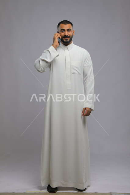 دردشات وتعارف باستخدام الهاتف المحمول, التواصل مع الأصدقاء بحب وود, بورتريه لرجل عربي خليجي سعودي يرتدي الثوب التقليدي يقوم بإجراء مكالمة هاتفية عبر الجوال, صورة لكامل الجسم , خلفية رمادية