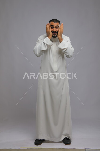 إزالة الكفين عن الوجه لرؤية المفاجأة, بورتريه لشاب عربي سعودي خليجي مبتسم يرتدي الثوب التقليدي ويقوم بتغطية وجهه للعب, مشاعر الملاعبة والملاطفة, قضاء وقت ممتع في التسلية والترفيه, صورة الجسم بالطول الكامل, خلفية رمادية.