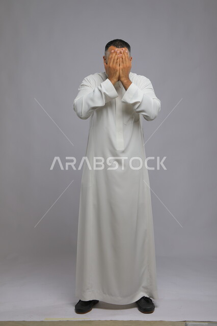 الاستعداد لرؤية المفاجأة, مفهوم اللعب و الملاطفة والمداعبه, بورتريه لشاب عربي خليجي سعودي يرتدي الثوب التقليدي يقوم بتغطية وجهه بكفيه, الشعور بالحزن والبكاء, صورة الجسم بالطول الكامل, خلفية رمادية.