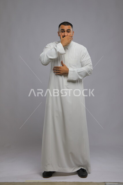الشعور بالسعادة والفرحة الشديدة, إيماءات الحياء والخجل, بورتريه لشاب عربي سعودي خليجي يرتدي الثوب التقليدي يضع يده على فمه ينظر الى الكاميرا , المفاجأة والدهشة عند سماع خبر ما, صورة الجسم بالطول الكامل, خلفية رمادية.