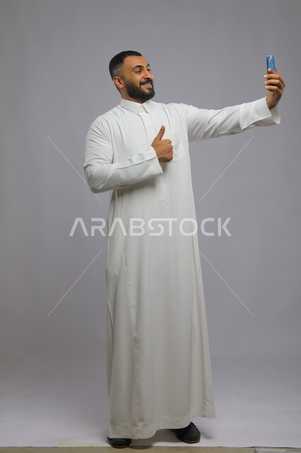 استخدام الأجهزة والتقنيات الحديثة, التقاط صور سيلفي تذكارية عبر الجوال, بورتريه لشاب عربي خليجي سعودي يرتدي الثوب التقليدي يقوم بحمل الهاتف المحمول, اجراء مكالمة فيديو عبر تطبيقات ومواقع السوشال ميديا, صورة الجسم بالكامل, خلفية رمادية