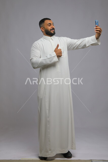 Taking souvenir selfies via mobile phone, using modern devices and technologies, a portrait of a Saudi Gulf Arab young man wearing a traditional dress holding a mobile phone, making a video call via social media applications and websites, a full-length photo of the body, gray background