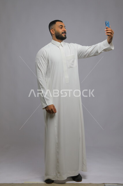 التقاط صور سيلفي تذكارية عبر الجوال, استخدام الأجهزة والتقنيات الحديثة, بورتريه لشاب عربي خليجي سعودي يرتدي الثوب التقليدي يقوم بحمل الهاتف المحمول, اجراء مكالمة فيديو عبر تطبيقات ومواقع السوشال ميديا, صورة الجسم بالطول الكامل, خلفية رمادية