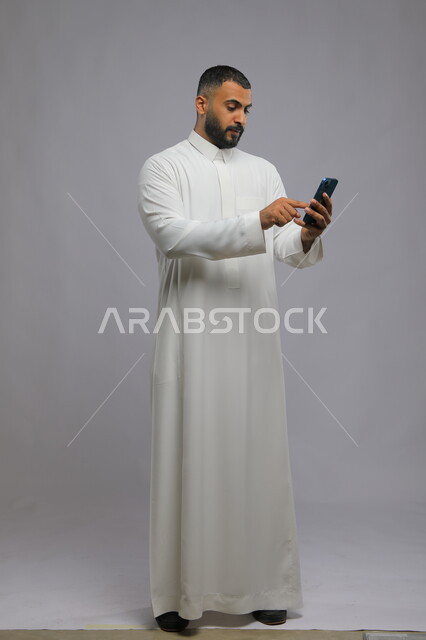 ايماءات النقر على الشاشة, بورتريه لشاب عربي خليجي سعودي يرتدي الثوب التقليدي يقوم باستخدام الهاتف المحمول, التسوق الالكتروني الذكي والآمن عبر الاونلاين, مفهوم صلة الرحم, صورة الجسم بالكامل, خلفية رمادية