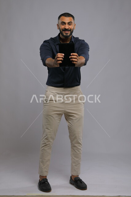 الاعجاب بتطبيقات وخدمات الجهاز اللوحي ،عرض شاشة اعلانية سوداء فارغة عبر التابليت، استخدام التقنيات الحديثة للتسويق الرقمي الالكتروني، بورتريه لشاب عربي خليجي سعودي مبتسم يرتدي الزي الكاجوال و ينظر إلى الكاميرا ،صورة الجسم بالطول الكامل، خلفية رمادية