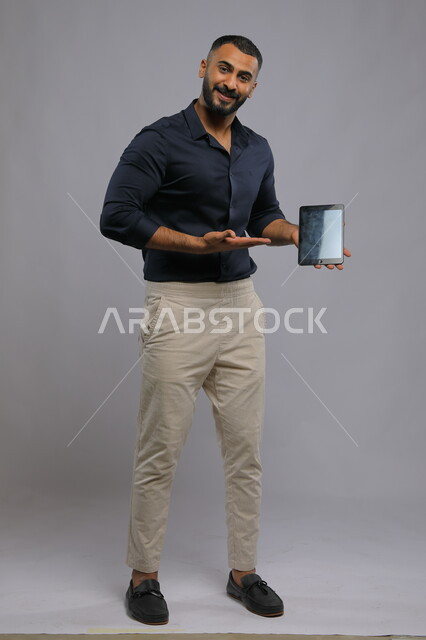 Gestures of admiration for the use of advanced modern technologies in practical life via tablet devices, portrait of a smiling Gulf Arab Saudi man wearing a casual outfit looking at the camera and pointing with his hand to the tablet with a blank black screen, full-length body photo, gray background