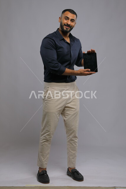 التسويق الإلكتروني عبر خدمات وتطبيقات الجهاز اللوحي، عرض شاشة سوداء فارغة عبر التابليت، بورتريه لشاب عربي خليجي سعودي مبتسم يرتدي الزي الكاجوال ينظر إلى الكاميرا بإيماءات السرور، دمج التقنية والتكنولوجيا في الحياة العملية، صورة الجسم بالطول الكامل، خلفية رمادية