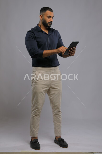تصفح الانترنت وإنجاز المهام عن بعد، متابعه آخر أخبار السوشال ميديا، عقد اجتماع عمل، استخدام التقنيات الحديثة المتطورة , بورتريه لرجل عربي خليجي سعودي يرتدي الزي الكاجوال يحمل بيده جهاز التابليت ،الدردشة مع الأهل والأصدقاء, صورة الجسم بالكامل, خلفية رمادية