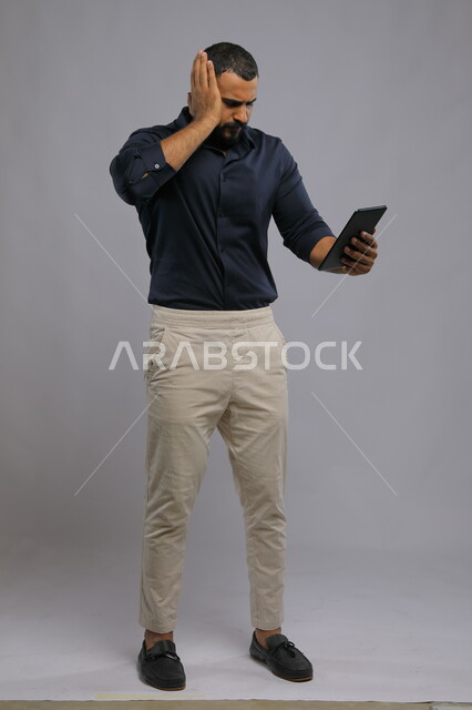 مشاهدة مباراة كرة القدم بإيماءات الغضب ,تشجييع المنتخب الوطني السعودي، استخدام التكنولوجيا و التقنيات الحديثة، بورتريه لرجل عربي خليجي سعودي يرتدي الزي الكاجوال يحمل جهاز التابليت و يضع يده على رأسه بتعابير الخسارة، صورة الجسم بالطول الكامل, خلفية رمادية