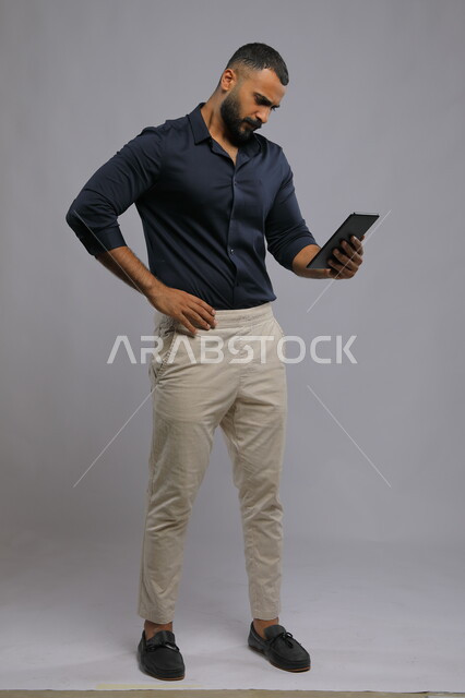 وضع اليد على الخصر بإيماءات التفكير ,قراءة خبر غير سار عبر مواقع الانترنت، استخدام التكنولوجيا و التقنيات الحديثة، بورتريه لرجل عربي خليجي سعودي يرتدي الزي الكاجوال يحمل بيده جهاز التابليت بإيماءات الانزعاج ،صورة الجسم بالطول الكامل, خلفية رمادية