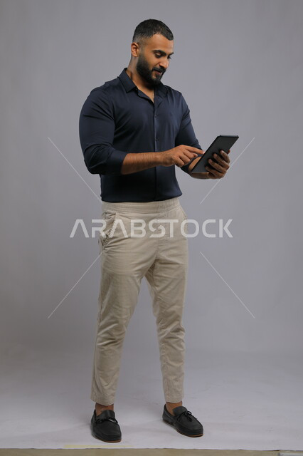  تطبيقات وبرامج التواصل الاجتماعي، متابعه آخر أخبار السوشال ميديا، عقد اجتماع عمل عن بعد، استخدام التقنيات الحديثة المتطورة , بورتريه لرجل عربي خليجي سعودي يرتدي الزي الكاجوال يحمل بيده جهاز التابليت ،الدردشة مع الأهل والأصدقاء, صورة الجسم بالكامل, خلفية رمادية