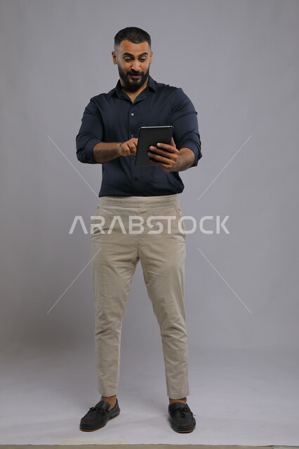  إيماءات السرور والاندماج أثناء اللعب على تطبيقات الأجهزة اللوحية, استخدام التقنيات الحديثة المتطورة , بورتريه لرجل عربي خليجي سعودي مبتسم يرتدي الزي الكاجوال يحمل بيده جهاز التابليت ،تعابير الفوز والنصر، صورة الجسم بالكامل, خلفية رمادية