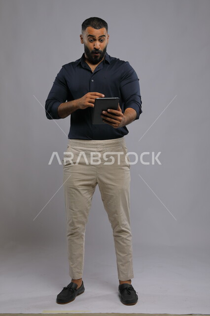Gestures of emotion and engagement while watching something on the tablet, enthusiasm and astonishment at victory and triumph, portrait of a Saudi Gulf Arab young man wearing casual clothes making a video call and hearing surprising news, full-length body portrait, gray background
