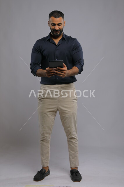 إجراء مكالمة فيديو و الدردشة مع الأهل بإيماءات السرور, استخدام التقنيات الحديثة المتطورة , بورتريه لرجل عربي خليجي سعودي مبتسم يرتدي الزي الكاجوال يحمل بيده جهاز التابليت ،متابعه آخر أخبار السوشال ميديا، تطبيقات الأجهزة اللوحية، صورة الجسم بالكامل, خلفية رمادية