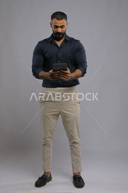 Watching favorite and entertaining programs, using advanced modern technologies, a portrait of a Saudi Gulf Arab man wearing a casual outfit holding a tablet in his hand, chatting with family and relatives, following the latest social media news, tablet applications, full body photo, gray background