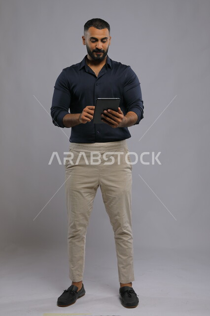 عقد اجتماع عمل عن بعد، استخدام التقنيات الحديثة المتطورة , بورتريه لرجل عربي خليجي سعودي يرتدي الزي الكاجوال يحمل بيده جهاز التابليت ،الدردشة مع الأهل والأصدقاء, متابعه آخر أخبار السوشال ميديا، تطبيقات وبرامج الأجهزة اللوحية، صورة الجسم بالكامل, خلفية رمادية