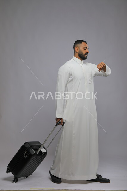 النظر إلى ساعه اليد للتاكد من الوقت، قضاء وقت ممتع في السياحه بالعطلة الصيفيه, التأخر على موعد الطائرة، بورتريه لشاب عربي خليجي سعودي يرتدي الثوب التقليدي يقوم بجر حقيبة سفر، صورة الجسم بالكامل, خلفية رمادية