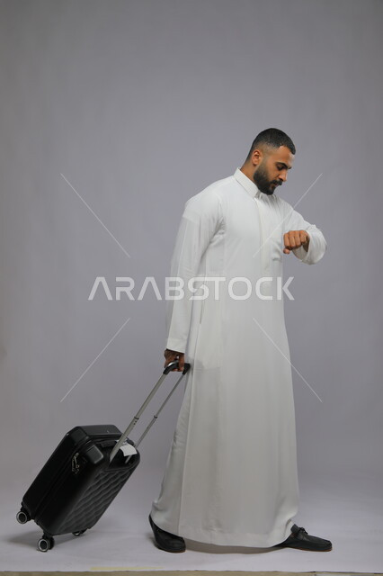النظر إلى ساعه اليد للتاكد من الوقت، قضاء وقت ممتع في السياحه بالعطلة الصيفيه, التأخر على موعد الطائرة، بورتريه لشاب عربي خليجي سعودي يرتدي الثوب التقليدي يقوم بجر حقيبة سفر، صورة الجسم بالكامل, خلفية رمادية