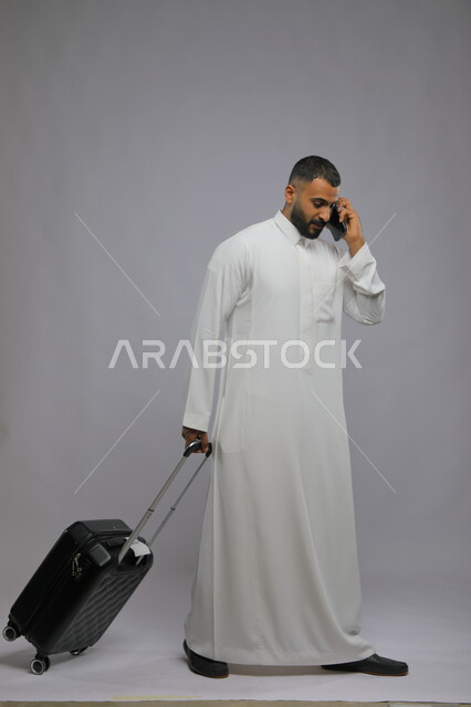 حجز مباشر عبر تطبيقات وبرامج السياحة, التواصل مع الأهل والأصدقاء, إجراء مكالمات هاتفية عبر الجوال, بورتريه لشاب عربي سعودي خليجي مبتسم يرتدي الثوب التقليدي يمسك بيده الهاتف المحمول وحقيبة السفر, صورة الجسم بالطول الكامل, خلفية رمادية