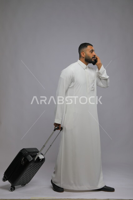 حجز مباشر عبر تطبيقات وبرامج السياحة, التواصل مع الأهل والأصدقاء, إجراء مكالمات هاتفية عبر الجوال, بورتريه لشاب عربي سعودي خليجي مبتسم يرتدي الثوب التقليدي يمسك بيده الهاتف المحمول وحقيبة السفر, صورة الجسم بالطول الكامل, خلفية رمادية