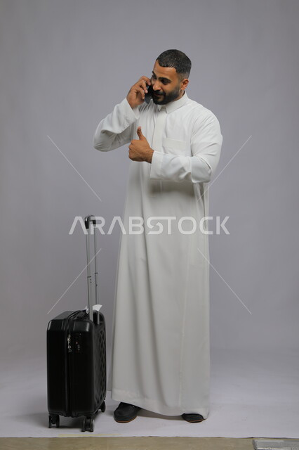 رفع اصبع الابهام نحو الأعلى بعلامة الجودة, بورتريه لرجل عربي سعودي خليجي يرتدي الثوب التقليدي يقوم بإجراء مكالمة هاتفية عبر الهاتف المحمول, التأكد من الحجز للرحلة عبر التقنيات الحديثة, صورة الجسم بالطول الكامل, خلفية رمادية