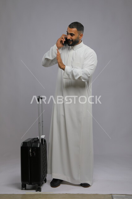 إجراء مكالمة هاتفية عبر الجوال, رفع اليد والتلويح للوداع والسلام, التواصل مع الأهل والأصدقاء, بورتريه لشاب عربي سعودي خليجي مبتسم يرتدي الثوب التقليدي يمسك الهاتف المحمول, حجز مباشر عبر تطبيقات وبرامج السياحة, صورة الجسم بالطول الكامل, خلفية رمادية