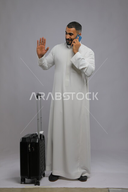 إجراء مكالمة هاتفية عبر الجوال, رفع اليد والتلويح للوداع والسلام, التواصل مع الأهل والأصدقاء, بورتريه لشاب عربي سعودي خليجي مبتسم يرتدي الثوب التقليدي يمسك الهاتف المحمول, حجز مباشر عبر تطبيقات وبرامج السياحة, صورة الجسم بالطول الكامل, خلفية رمادية