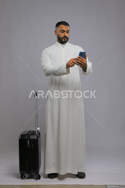 إرسال رسالة نصية عبر الجوال لتأكيد الحجز, قضاء أوقات ممتعه في السياحة في العطلة الصيفية, بورتريه لشاب عربي خليجي سعودي يرتدي الثوب التقليدي يقوم باستخدام الهاتف المحمول, تصفح مواقع التواصل الاجتماعي, صورة الجسم بالطول الكامل, خلفية رمادية