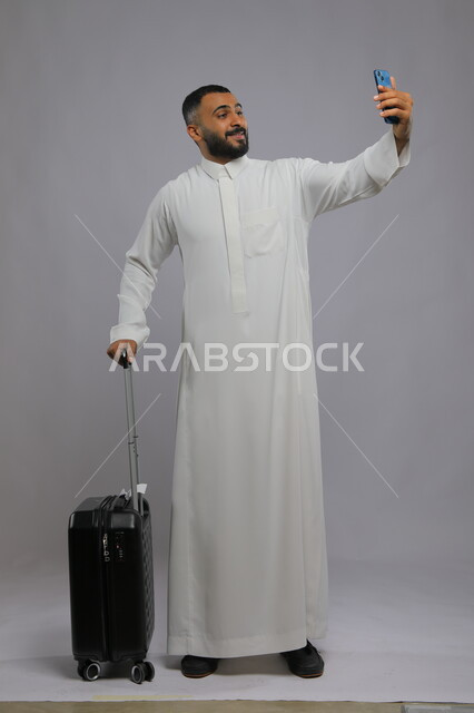 التقاط صور سيلفي تذكارية لتوثيق اللحظات السعيدة, مفهوم السياحة والسفر, بورتريه لشاب عربي خليجي سعودي يرتدي الثوب التقليدي يمسك حقيبة السفر والهاتف المحمول بإيماءات الحماس والسرور, صورة الجسم بالطول الكامل, خلفية رمادية
