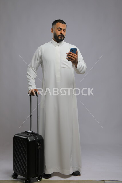 مشاهدة البرامج المفضلة عبر الجوال, قضاء أوقات ممتعه في السياحة في العطلة الصيفية, بورتريه لشاب عربي خليجي سعودي يرتدي الثوب التقليدي يقوم بحمل حقيبة السفر والهاتف المحمول, تصفح مواقع التواصل الاجتماعي, صورة الجسم بالطول الكامل, خلفية رمادية